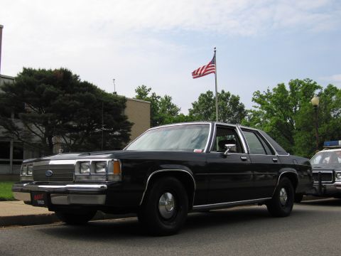 1991 Ford LTD Crown Victoria - black Ford LTD Crown Victoria sedan parked at a curb near a U.S. flag, with another police cruiser visible at right.