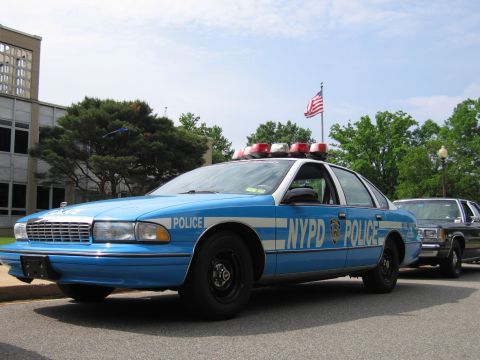 1991 Ford LTD Crown Victoria - blue-and-white NYPD Chevrolet Caprice police car with roof lights and door lettering parked beside a building near a flag.