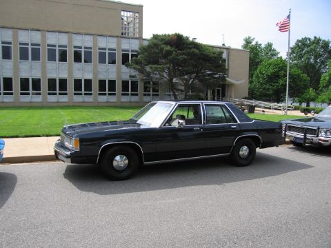 1991 Ford LTD Crown Victoria - black four-door full-size sedan parked at a curb in front of a tan office building, with an American flag and another car nearby.