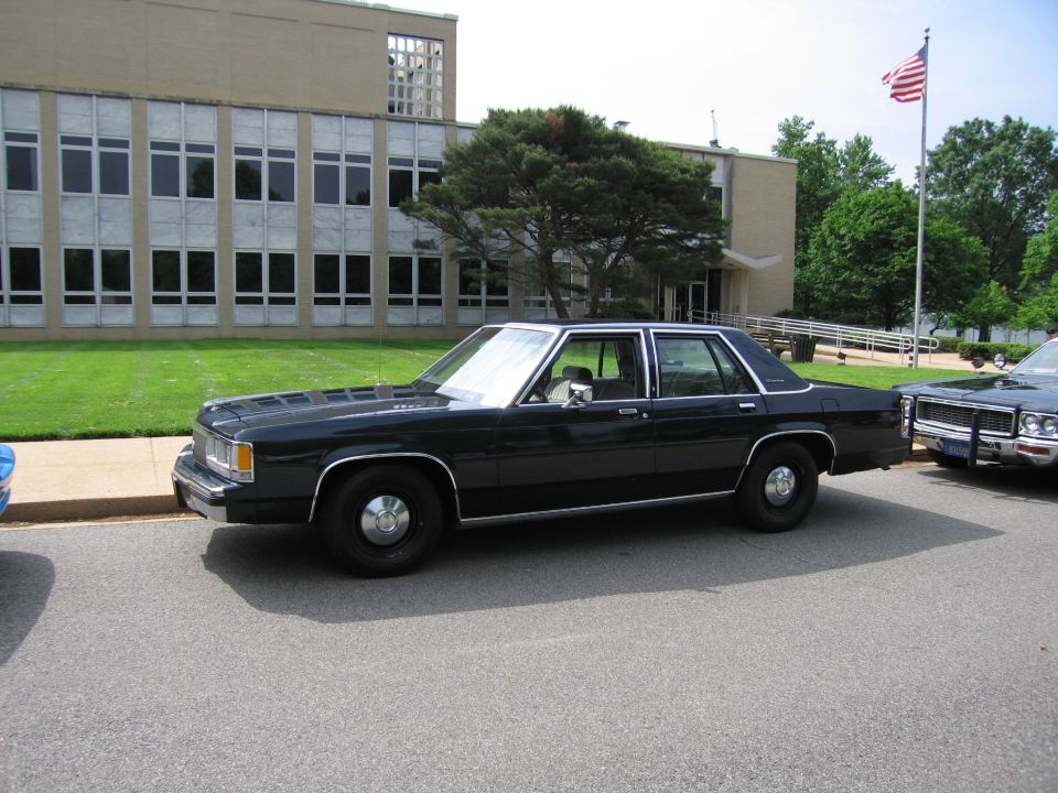 Black Ford LTD Crown Victoria-style sedan parked by a building with US flag