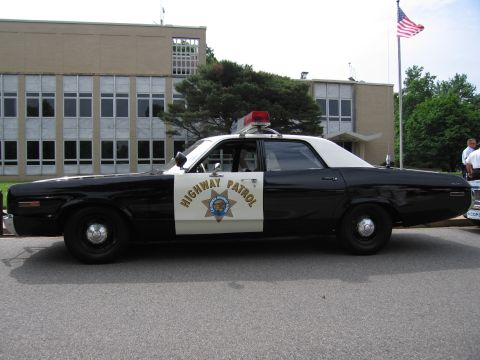 1991 Ford LTD Crown Victoria - black-and-white police sedan lettered HIGHWAY PATROL with a gold star emblem, parked before a tan building flying an American flag.