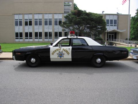 1991 Ford LTD Crown Victoria - Side profile of a black-and-white HIGHWAY PATROL cruiser with a red roof light, parked in front of a building with large windows.