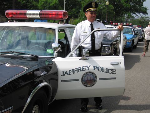 1991 Ford LTD Crown Victoria - uniformed officer stands holding open the white door of a JAFFREY POLICE cruiser, with a row of other police cars behind.