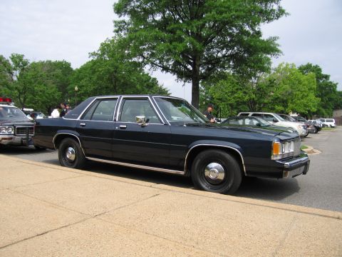 1991 Ford LTD Crown Victoria - dark navy four-door full-size sedan parked along a curb in a tree-lined lot with other vehicles and people behind.
