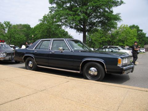 1991 Ford LTD Crown Victoria - dark navy four-door sedan parked at a curb while a uniformed officer stands near its right rear, other cars in the lot behind.