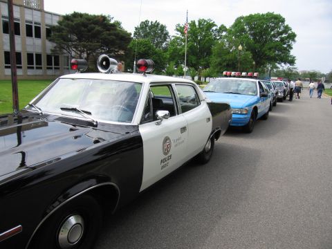 1991 Ford LTD Crown Victoria - black-and-white police sedan with twin roof lights and POLICE lettering leads a long row of parked cruisers along a drive.