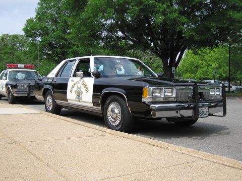 1991 Ford LTD Crown Victoria - Front three-quarter view of a black-and-white HIGHWAY PATROL sedan with push bar and star door emblem parked beside another cruiser.