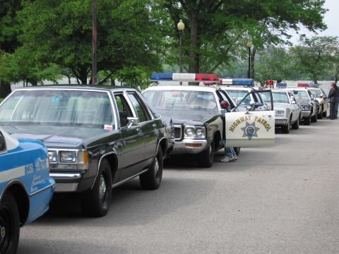 1991 Ford LTD Crown Victoria - long row of parked vintage police cruisers, including a black-and-white Highway Patrol car and a blue cruiser, lined along a curb.