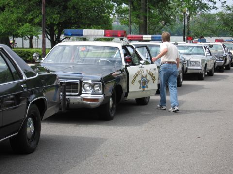 1991 Ford LTD Crown Victoria - man stands at the open door of a black-and-white HIGHWAY PATROL sedan in a long lineup of police cars beside water and trees.