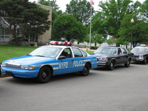 1991 Ford LTD Crown Victoria - blue and white NYPD POLICE Chevrolet Caprice cruiser parked at a curb ahead of two black sedans, with a building and flag behind.