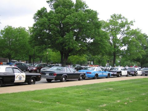 1991 Ford LTD Crown Victoria - lineup of classic police cruisers, including a blue NYPD-marked car, parked along a grassy tree-shaded street.