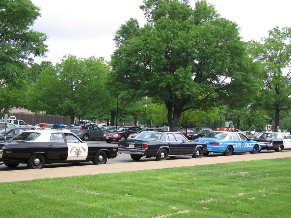 Classic police cars lined up on a park road, including a black-and-white cruiser and a blue NYPD sedan