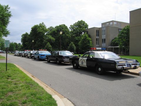 1991 Ford LTD Crown Victoria - black-and-white Highway Patrol cruiser parked at the front of a row of vintage police cars beside a brick building.