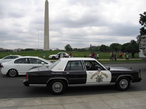 1991 Ford LTD Crown Victoria - black-and-white Highway Patrol cruiser parked beside a white car, the Washington Monument and a crowd in the background.