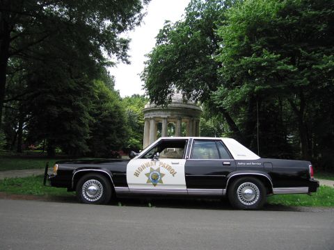 1991 Ford LTD Crown Victoria - black-and-white Highway Patrol cruiser parked beside a grassy path with a domed columned gazebo among trees.