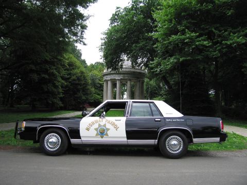 1991 Ford LTD Crown Victoria - Driver-side view of a black-and-white Highway Patrol cruiser parked before a domed columned stone gazebo in a park.