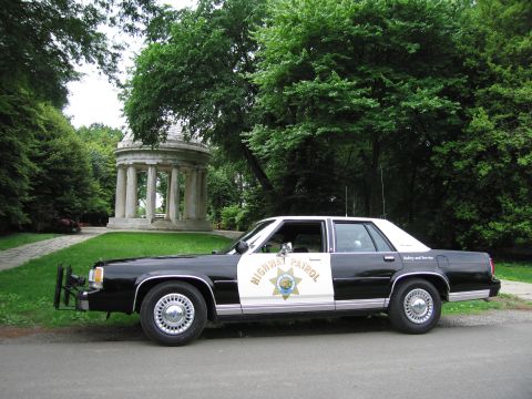 1991 Ford LTD Crown Victoria - black-and-white Highway Patrol sedan with a star door emblem sits on a tree-lined park path beside a classical domed monument.