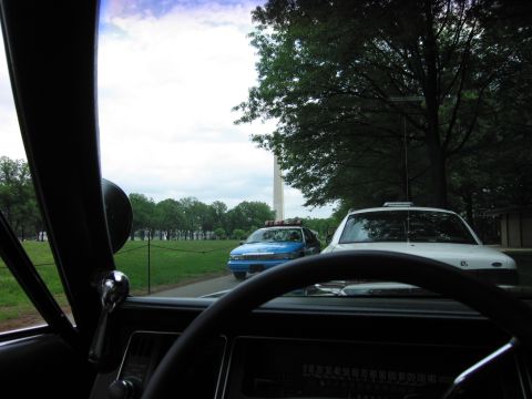 1991 Ford LTD Crown Victoria - From inside a car, a blue-and-white police car and a white sedan sit on a path near the Washington Monument under a cloudy sky.