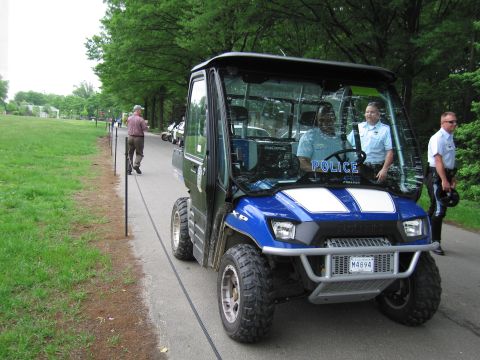 1991 Ford LTD Crown Victoria - Officers stand beside a blue-and-black Polaris police utility vehicle marked POLICE on a paved path through a park.