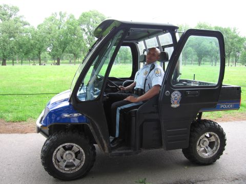 1991 Ford LTD Crown Victoria - Side profile of a blue-and-black Polaris police side-by-side with a uniformed officer in the driver's seat on a park path.