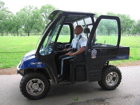 1991 Ford LTD Crown Victoria - uniformed officer drives a blue-and-black Polaris police utility vehicle along a paved path beside an open grassy field.