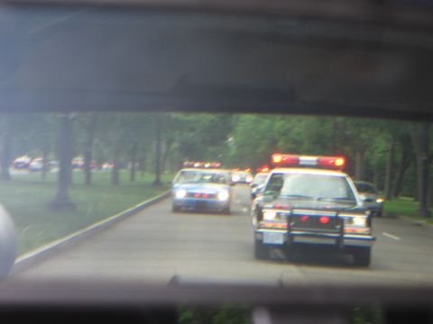 1991 Ford LTD Crown Victoria - slightly blurred windshield view of police cars with lit red roof bars driving in a column beneath a highway overpass.