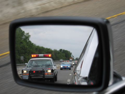 1991 Ford LTD Crown Victoria - car's side mirror reflects a black police cruiser with a lit roof light bar following on a divided highway.