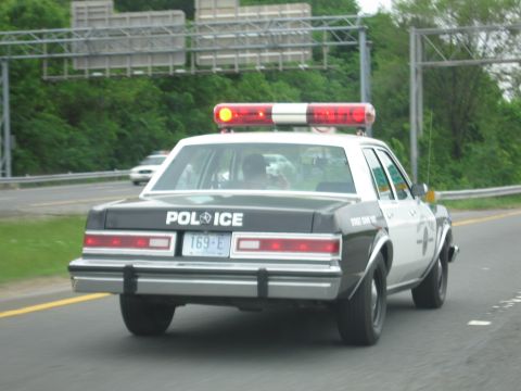 1991 Ford LTD Crown Victoria - black-and-white police sedan marked POLICE with a lit roof light bar drives along a multi-lane highway past overhead sign gantries.