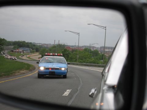 1991 Ford LTD Crown Victoria - car's side mirror reflects an oncoming blue police car with headlights and roof lights on a highway near a cityscape.
