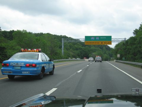 1991 Ford LTD Crown Victoria - blue-and-white police car with lit roof lights drives on a divided highway beneath green I-495/95 South signage reading Drawbridge 4 Miles.