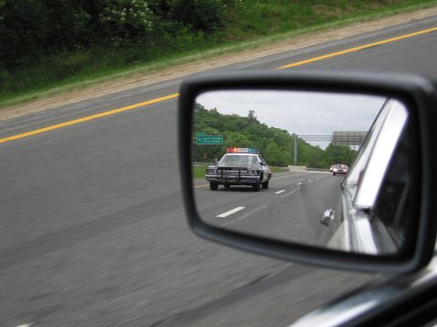 1991 Ford LTD Crown Victoria - car's side mirror reflects a black-and-white police cruiser with a lit roof bar following on a green highway shoulder.