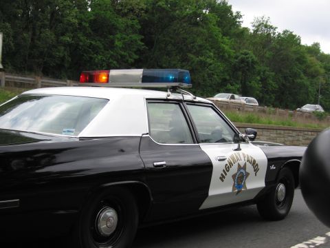 1991 Ford LTD Crown Victoria - black-and-white Highway Patrol cruiser with red and blue roof lights and a gold star shield travels along a tree-lined highway.