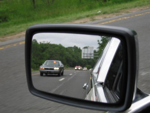 1991 Ford LTD Crown Victoria - car's side mirror reflects a dark vintage sedan with amber roof lights following on a tree-lined divided highway.