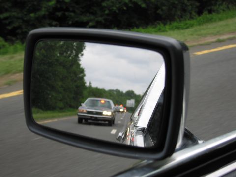 1991 Ford LTD Crown Victoria - side mirror reflects a dark sedan with amber roof lights and headlights on, following on a rural two-lane road.