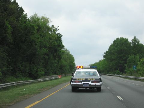 1991 Ford LTD Crown Victoria - Seen from behind, a black-and-white Highway Patrol sedan with roof lights drives ahead on a tree-lined highway under a cloudy sky.
