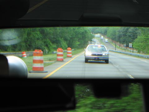 1991 Ford LTD Crown Victoria - Through a windshield, a patrol car with headlights on travels a rural highway past orange traffic barrels.