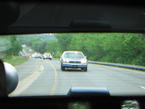 1991 Ford LTD Crown Victoria - Through a windshield, a line of vintage cars with headlights on travels along a curving tree-lined highway.