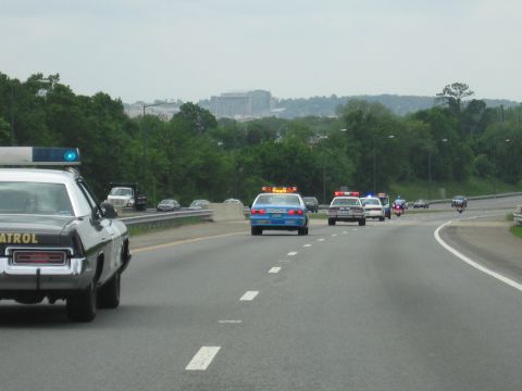 1991 Ford LTD Crown Victoria - convoy of vintage patrol cars and motorcycles with emergency lights travels a multi-lane highway with a distant skyline.