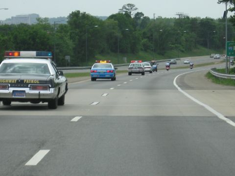 1991 Ford LTD Crown Victoria - black Highway Patrol sedan leads a procession of vintage cars and motorcycles along a tree-lined divided highway.