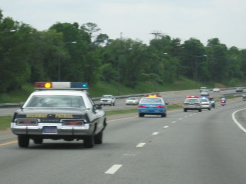 1991 Ford LTD Crown Victoria - black-and-white Highway Patrol sedan with amber and blue roof lights leads vintage cars and motorcycles along a highway.