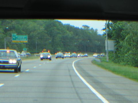 1991 Ford LTD Crown Victoria - Through a windshield, a long convoy of vintage cars travels a curving highway past a green directional sign.