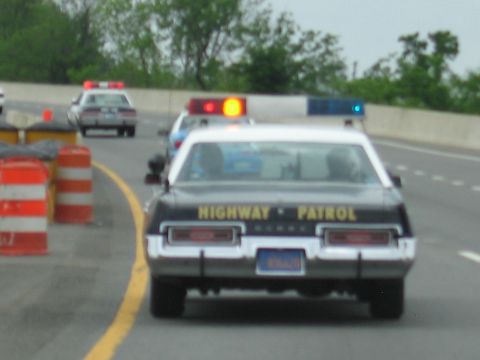 1991 Ford LTD Crown Victoria - rear of a black-and-white Highway Patrol sedan reading HIGHWAY PATROL shows roof lights as it passes orange barrels on a highway.