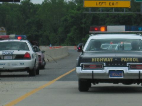 1991 Ford LTD Crown Victoria - white Metropolitan Police cruiser and a black Highway Patrol Dodge sit side by side on a highway beneath a yellow Left Exit sign.