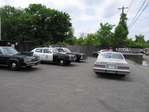 1991 Ford LTD Crown Victoria - row of vintage police cars, one marked STATE POLICE, sits parked in a gravel lot near a Save More used auto parts sign.