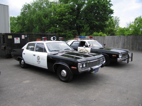 1991 Ford LTD Crown Victoria - black-and-white AMC marked POLICE sits beside a black Dodge patrol car on pavement near a wood fence and dumpster.