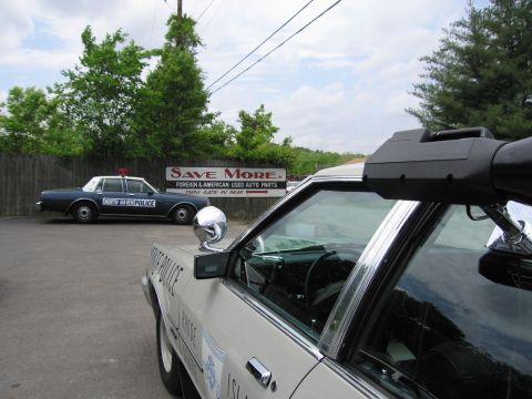 1991 Ford LTD Crown Victoria - white patrol car's door-mounted spotlight is in close view, with a North Haven Police sedan parked behind near a Save More sign.