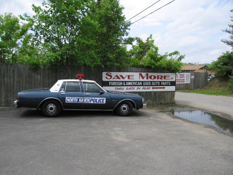 1991 Ford LTD Crown Victoria - dark blue North Haven Police sedan is parked in a gravel lot in front of a large Save More foreign and American used auto parts sign.