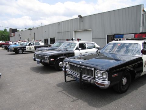 1991 Ford LTD Crown Victoria - row of vintage black-and-white police cars, including a Dodge with a push bar, sits parked at an angle outside a metal warehouse.