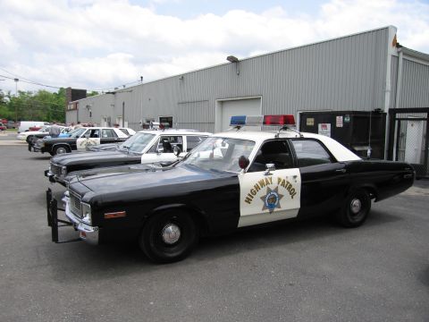 1991 Ford LTD Crown Victoria - black-and-white Dodge Highway Patrol sedan with a gold star shield is parked at an angle outside a metal warehouse among other cruisers.