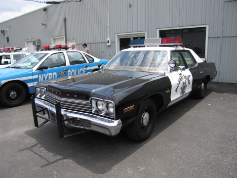 1991 Ford LTD Crown Victoria - black-and-white Dodge patrol car with a chrome push bar is parked beside a blue-striped NYPD cruiser outside a metal warehouse.